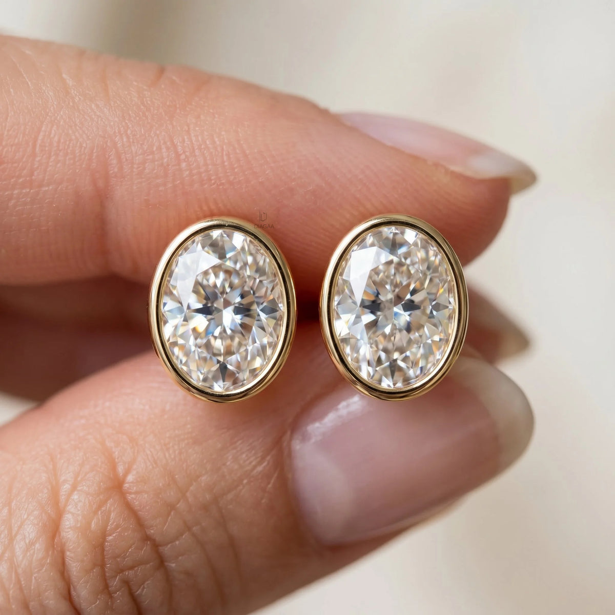 Close-up of fingers holding a pair of large yellow gold bezel-set oval diamond stud earrings to show scale and brilliance earring.