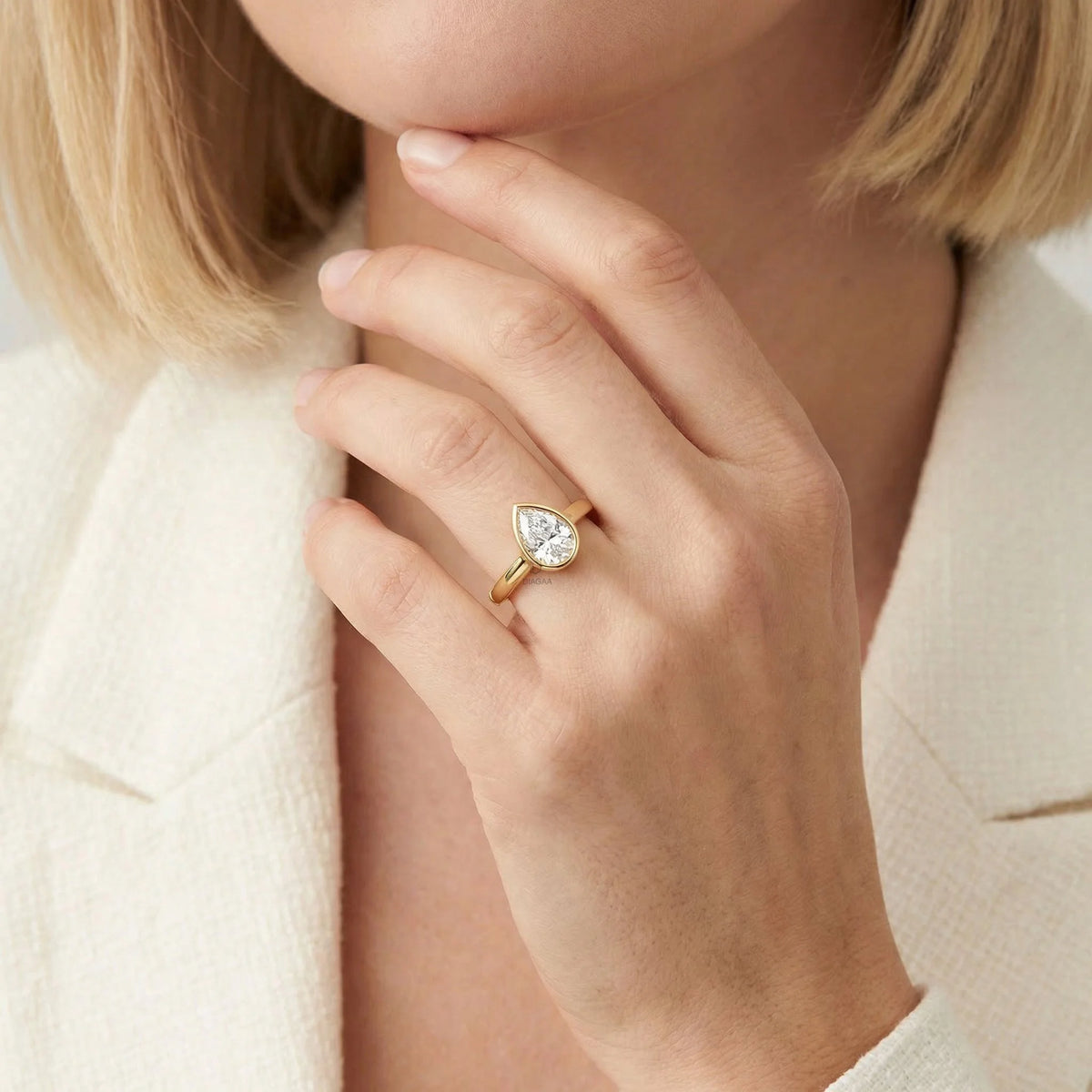 Close-up of a woman in a white blazer wearing a yellow gold engagement ring featuring a pear-shaped diamond in a modern bezel setting.