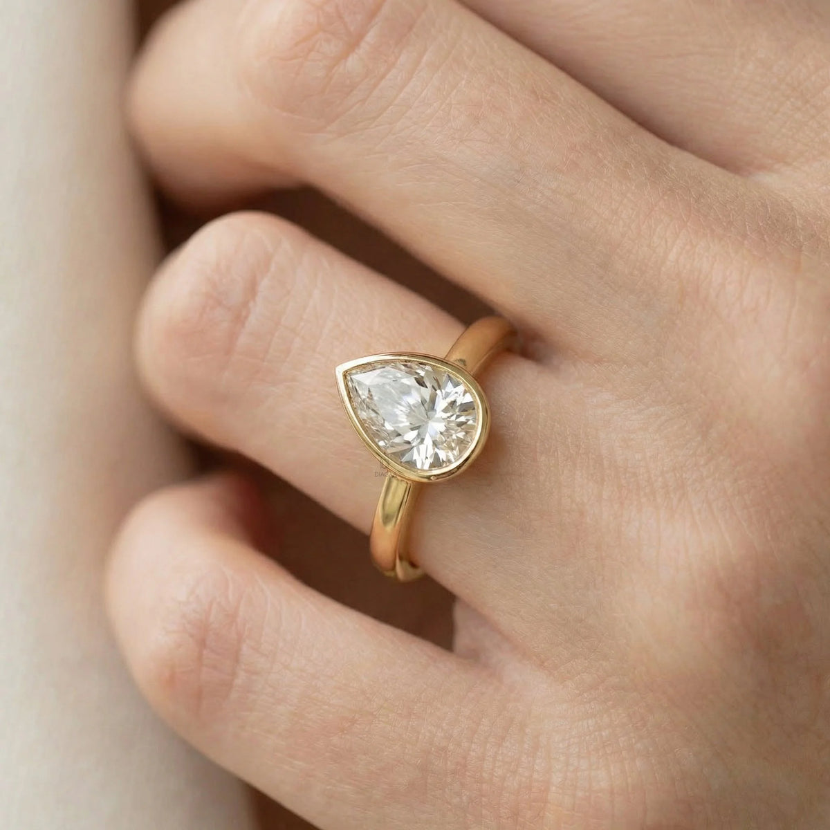 Close-up of a yellow gold pear-cut diamond engagement ring in a bezel setting, styled on a woman's hand 