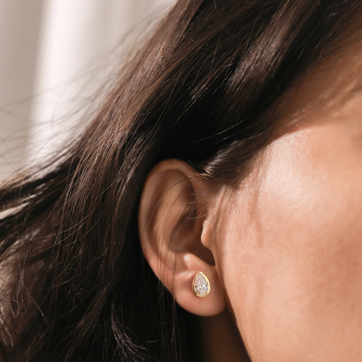 Woman wearing a gold bezel teardrop diamond stud earring. Dainty pear-shaped gemstone stud for ear lobe piercing, elegant fine jewelry