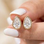 Close-up of fingers holding a pair of bezel-set pear-cut diamond stud earrings in yellow gold. Elegant teardrop fine jewelry.