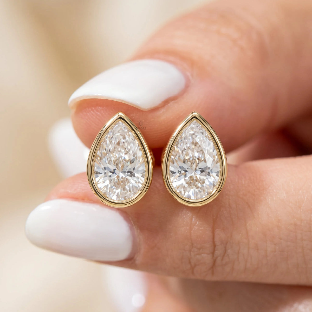 Close-up of fingers holding a pair of bezel-set pear-cut diamond stud earrings in yellow gold. Elegant teardrop fine jewelry.