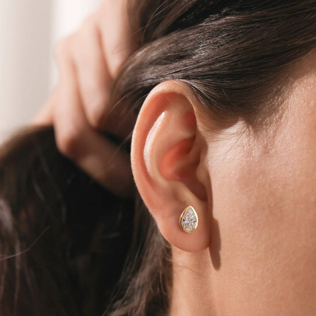 Woman wearing a minimalist pear cut diamond stud earring featuring a clear teardrop gemstone in a smooth gold bezel setting earring.