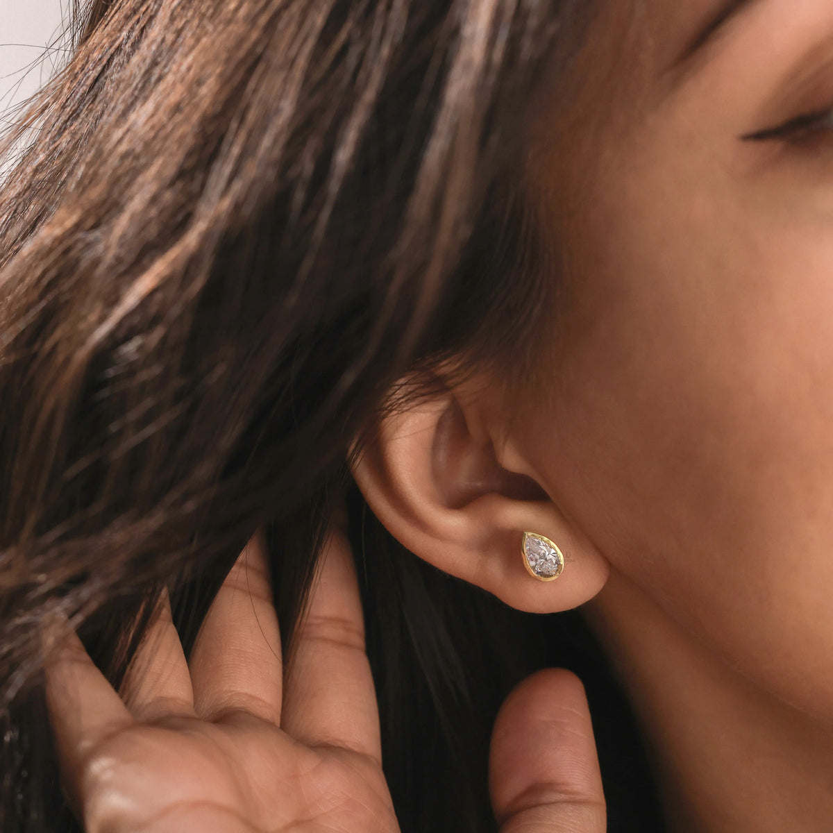 Side profile portrait of a woman showcasing a yellow gold pear cut diamond stud earring set in a smooth bezel frame. Modern daily jewelry.