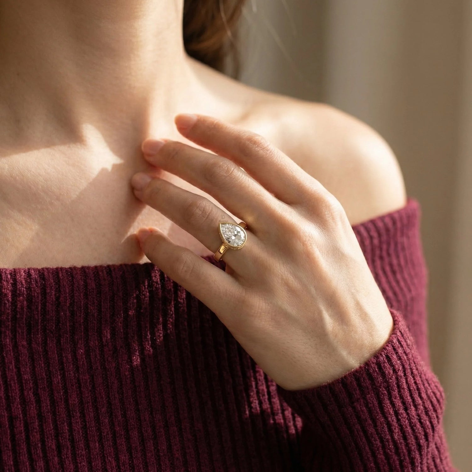 Close-up of a hand wearing a stunning pear-cut diamond ring in a yellow gold bezel setting, paired with a maroon knit sweater under warm sunlight