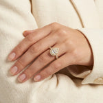 Yellow gold engagement ring featuring a pear shaped center diamond with a sparkling halo and pave band, modeled on a hand against a cream blazer.