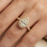 A pear shaped diamond engagement ring with a sparkling diamond halo and a pavé accented yellow gold band being worn on a finger.