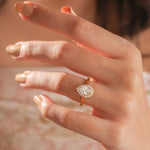 Hand wearing a gold pear diamond ring against . This unique Solitaire pear diamond ring showcases a bright center stone band. round diamond halo set ring.