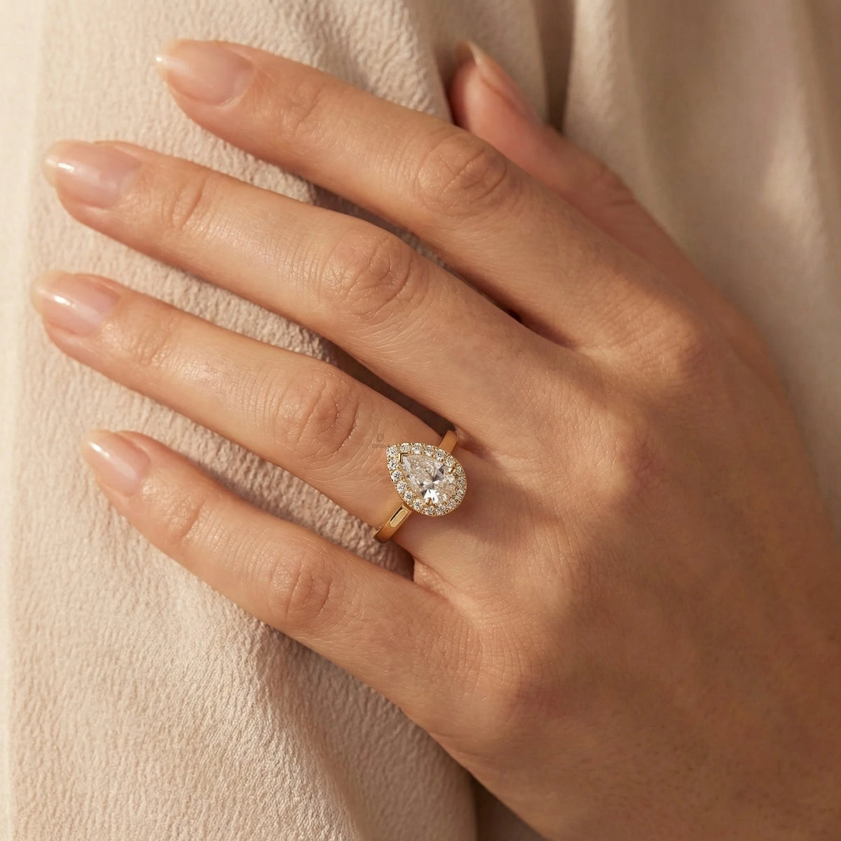 Hand wearing a 14k yellow gold pear diamond ring against . This unique Solitaire pear diamond ring showcases a bright center stone band. round diamond halo set ring.