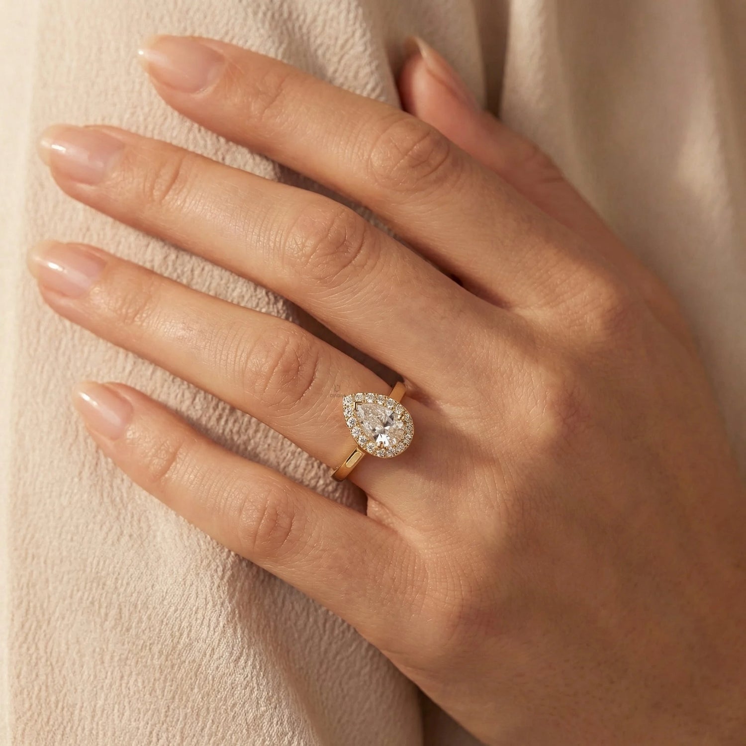 Hand wearing a 14k yellow gold pear diamond ring against . This unique Solitaire pear diamond ring showcases a bright center stone band. round diamond halo set ring.