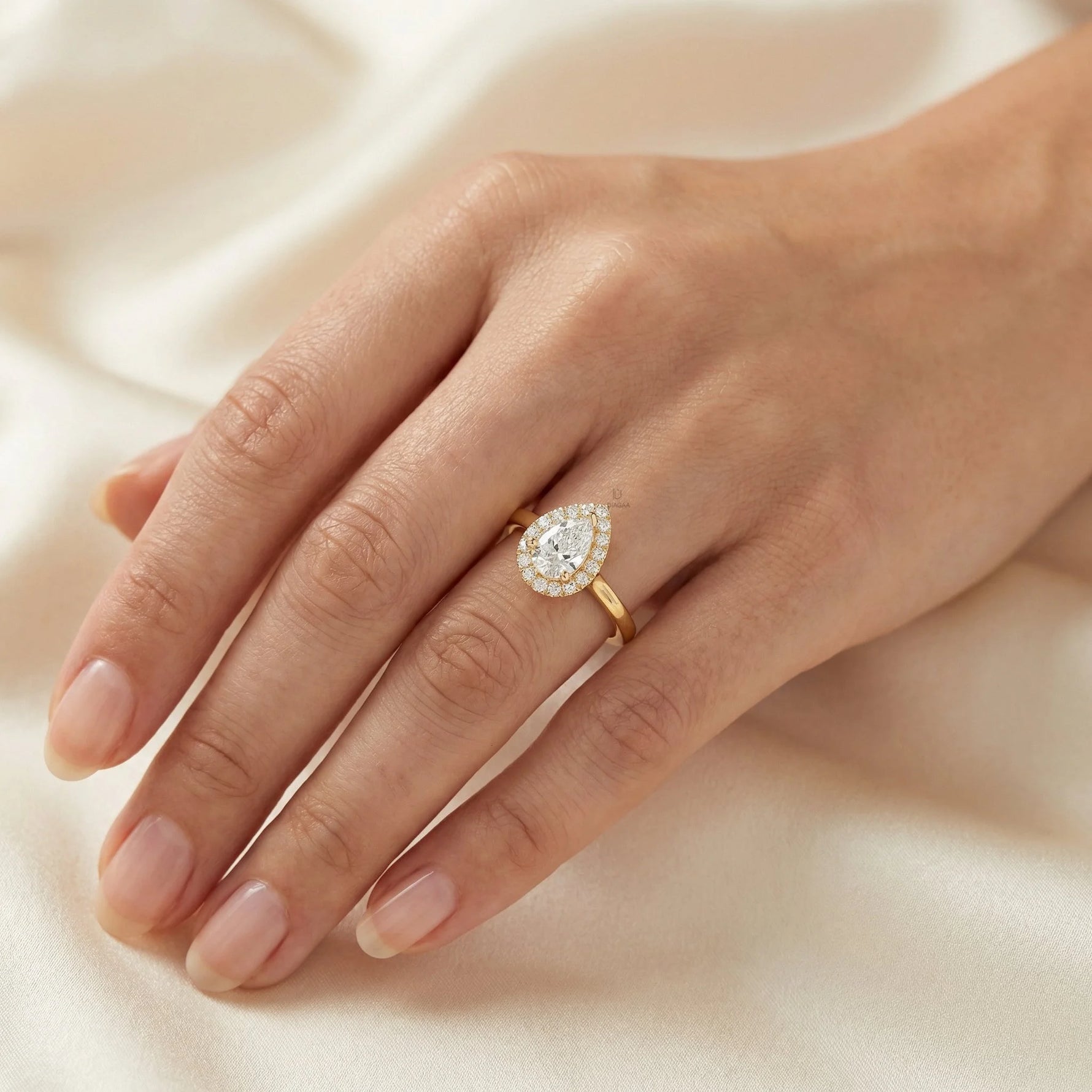 Hand wearing a yellow gold pear diamond ring against . This unique Solitaire pear diamond ring showcases a bright center stone band. round diamond halo set ring.