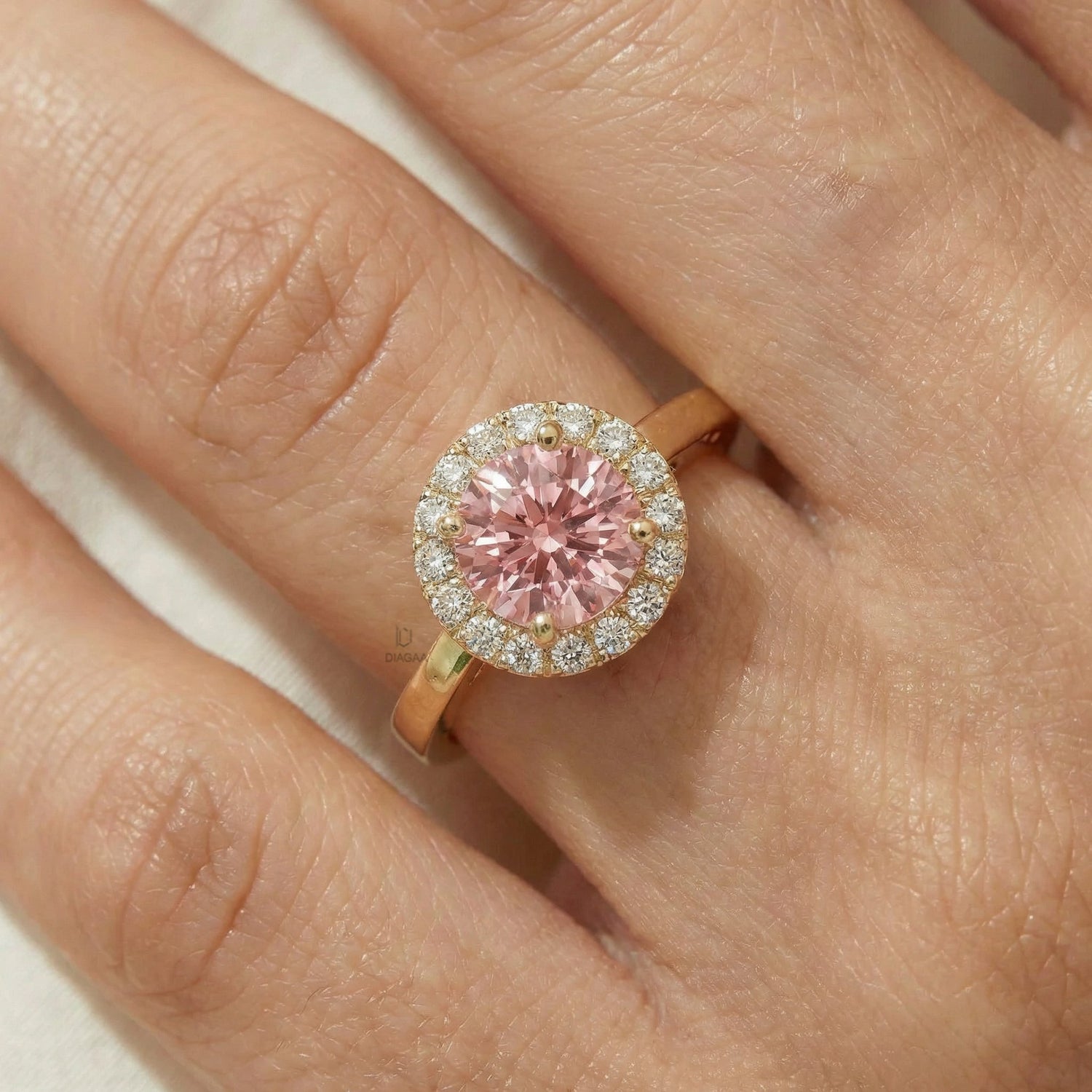 Close-up of a hand wearing a pink halo engagement ring in yellow gold, featuring a round pink center stone and diamond halo on soft cream silk.