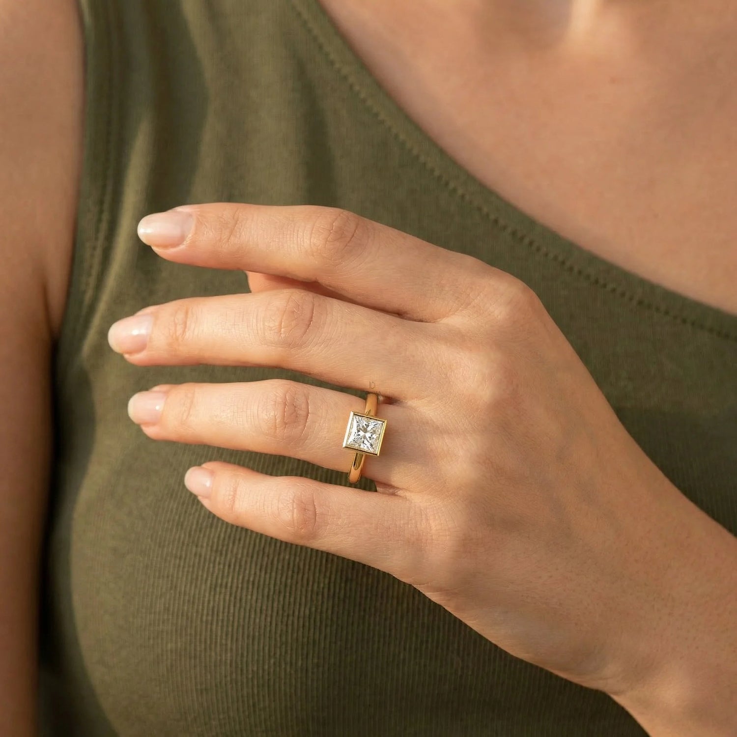 Close-up of a yellow gold princess-cut diamond engagement ring in a bezel setting, styled on a woman's hand wearing an olive green sleeveless top.