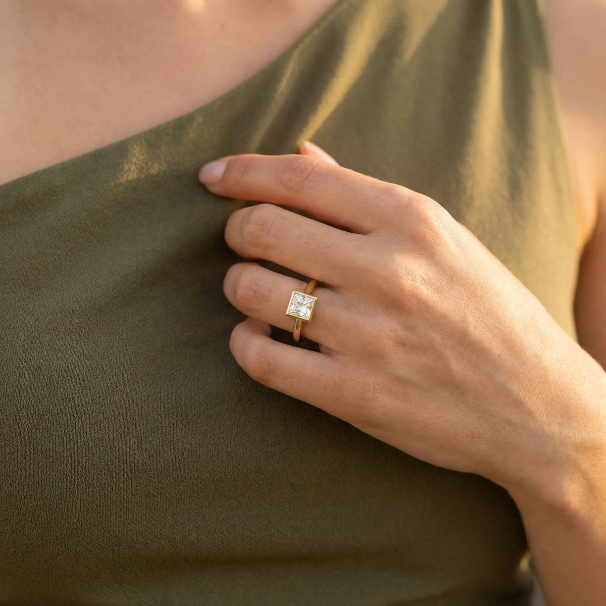Minimalist yellow gold bezel solitaire ring featuring a square princess diamond, displayed gracefully on a woman’s ring finger for a clean bridal look.