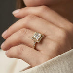Woman's hand wearing a yellow gold princess halo diamond engagement ring with a pave band, resting on a dark surface while wearing a cream blazer.
