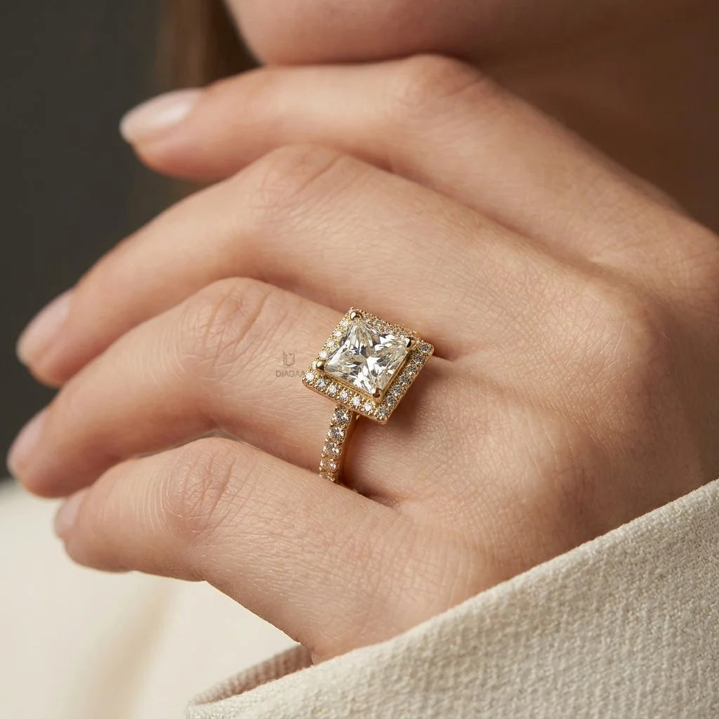 Woman's hand wearing a yellow gold princess halo diamond engagement ring with a pave band, resting on a dark surface while wearing a cream blazer.