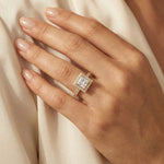 Close-up of a woman's hand resting on cream silk, wearing a yellow gold engagement ring with a princess-cut diamond, square halo, and pave band.