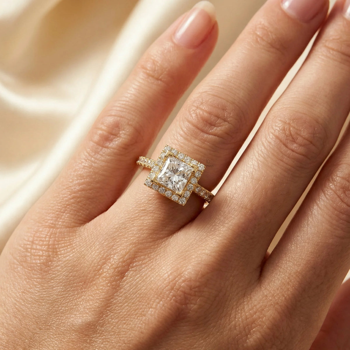 Woman's hand wearing a yellow gold princess halo diamond engagement ring with a pave band, resting on a dark surface while wearing a cream blazer.