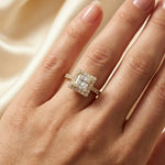 Woman's hand wearing a yellow gold princess halo diamond engagement ring with a pave band, resting on a dark surface while wearing a cream blazer.