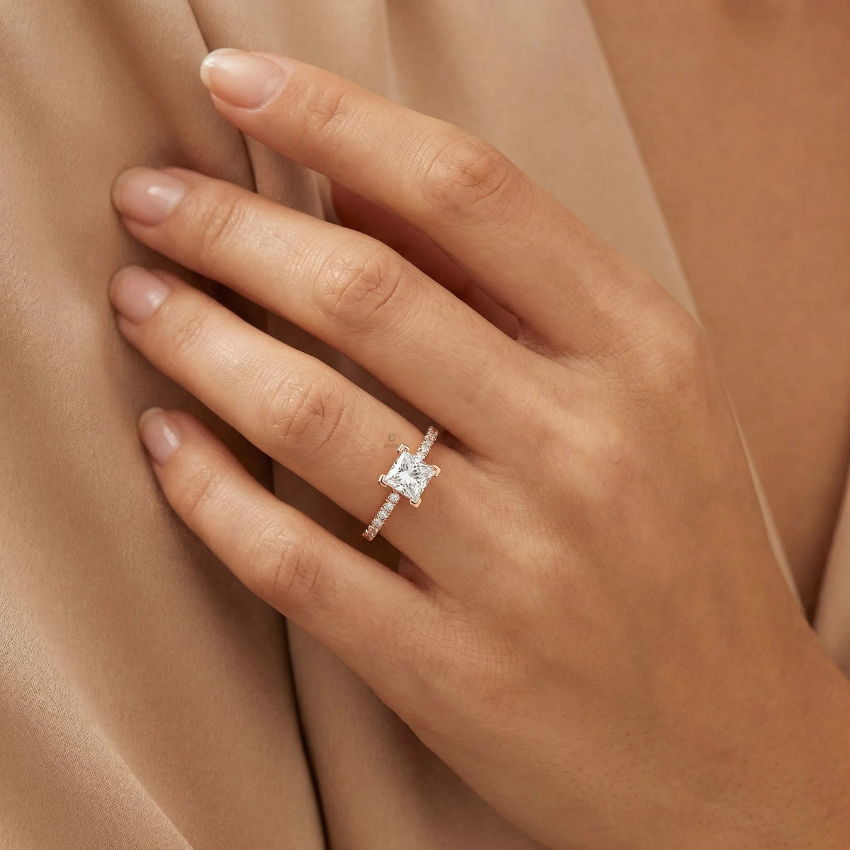 Woman's hand wearing a yellow gold engagement ring with a princess cut center diamond and round cut pavé band against soft beige fabric.