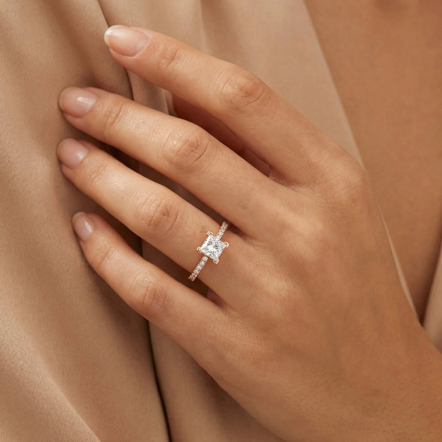Woman's hand wearing a yellow gold engagement ring with a princess cut center diamond and round cut pavé band against soft beige fabric.