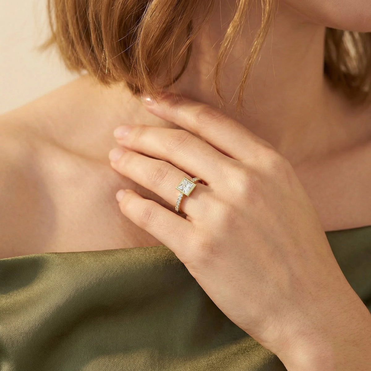 Close-up of a 10k yellow gold princess-cut diamond engagement ring with a modern bezel setting and shimmering pavé band, styled on a woman's hand wearing an olive green top.