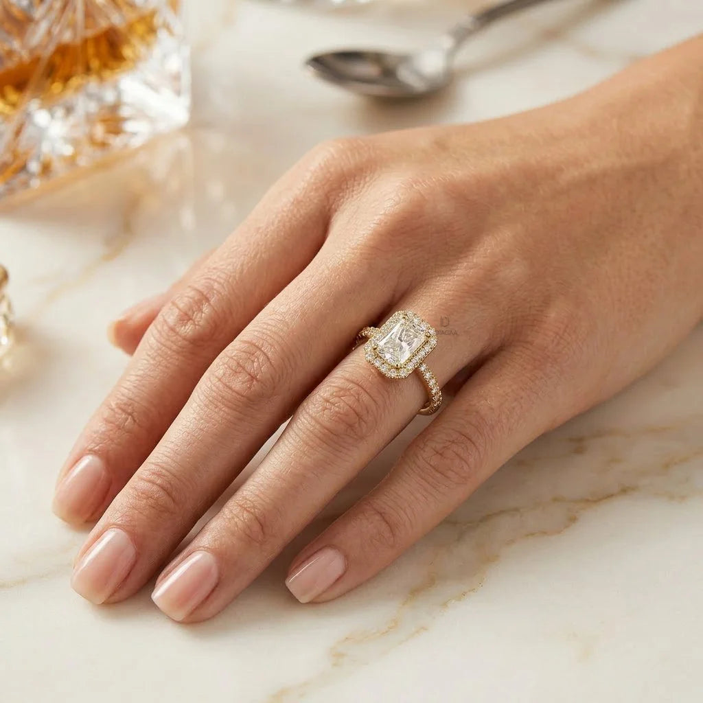 Woman's hand resting on white marble, wearing a yellow gold engagement ring featuring a radiant-cut diamond with a round cut halo and pave set band.