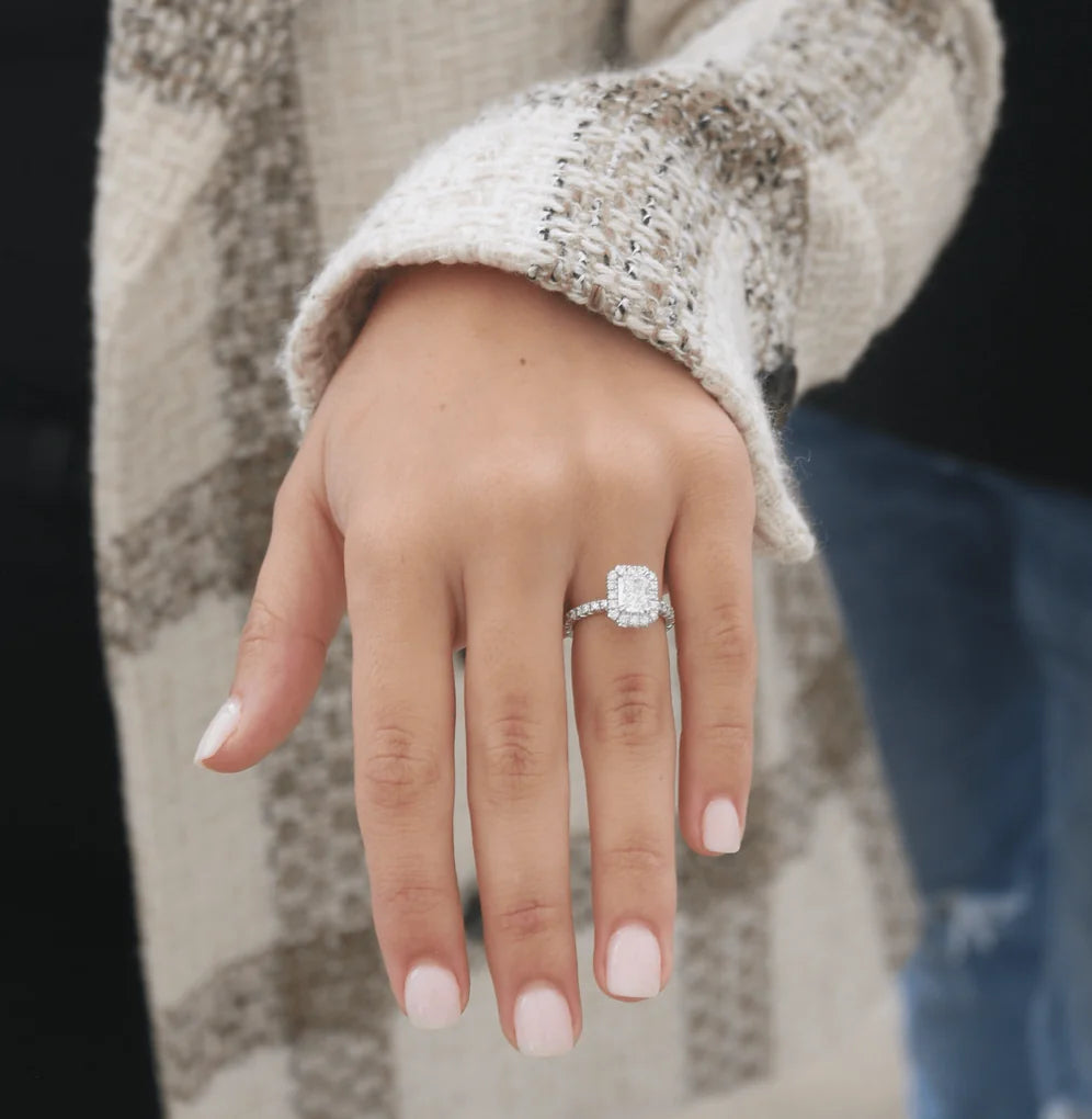 Elegant yellow gold engagement ring showcasing a rectangular center diamond, sparkling halo, and pave band, worn on a hand against a light marble surface.