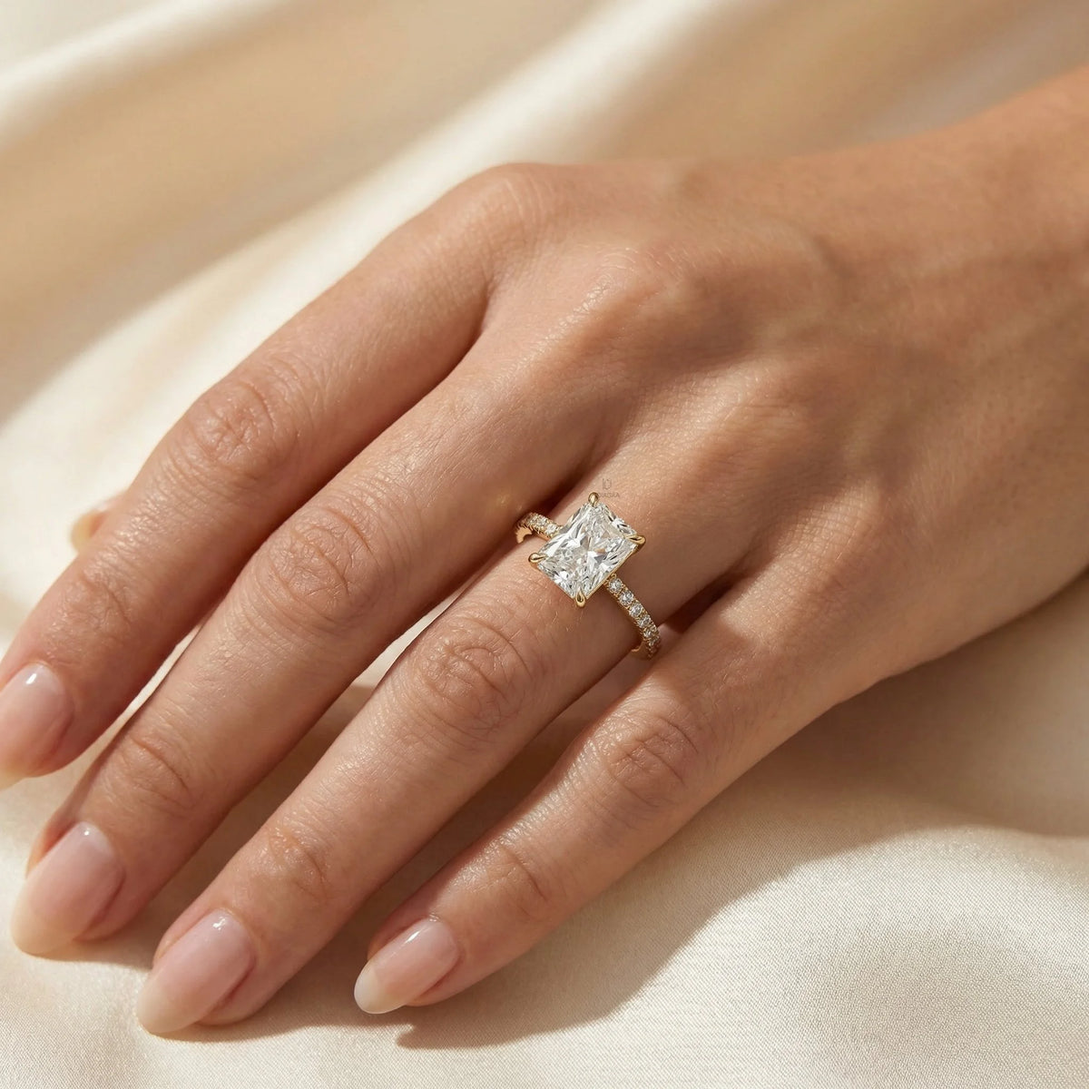 Woman's hand wearing a 14k yellow gold radiant cut diamond engagement ring with a round diamond pavé band on beige silk background.