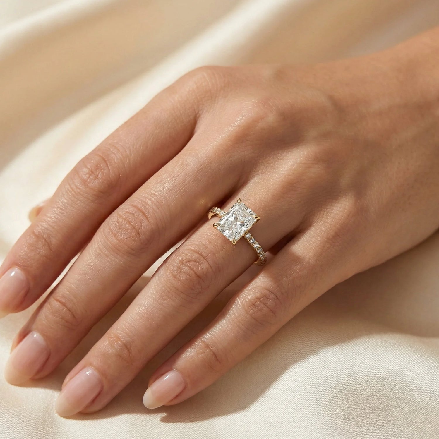 Woman's hand wearing a 14k yellow gold radiant cut diamond engagement ring with a round diamond pavé band on beige silk background.