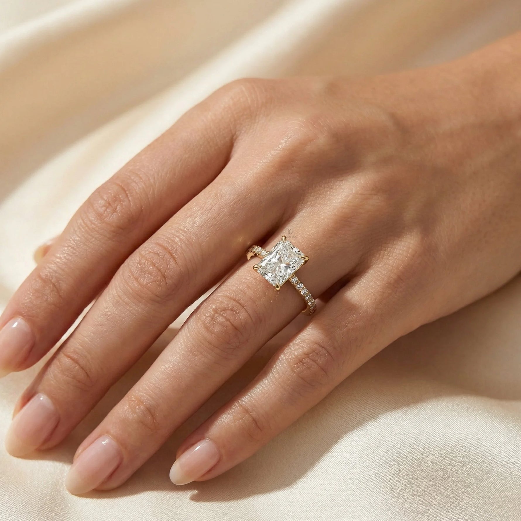 Woman's hand wearing a 14k yellow gold radiant cut diamond engagement ring with a round diamond pavé band on beige silk background.