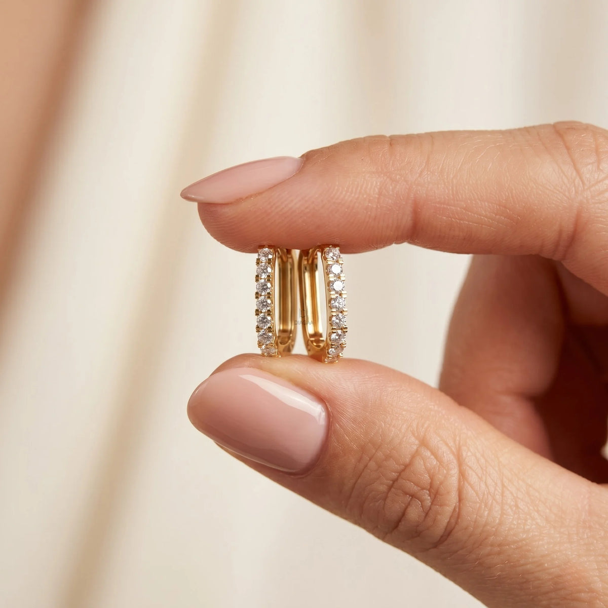 Fingers holding small yellow gold round cut rectangular huggie hoop earrings lined with sparkling diamonds against a soft beige background.