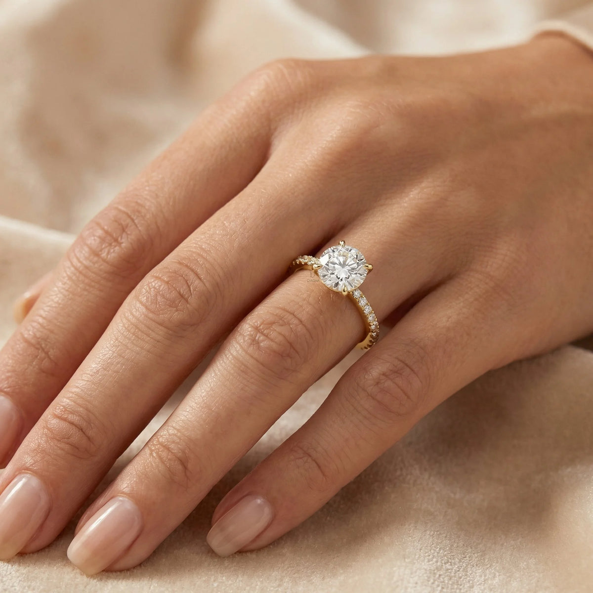 Round cut diamond engagement ring on a yellow gold side stone pavé set band worn on a woman's hand against a soft beige fabric.