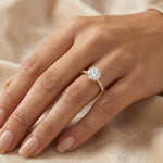 Round cut diamond engagement ring on a yellow gold side stone pavé set band worn on a woman's hand against a soft beige fabric.