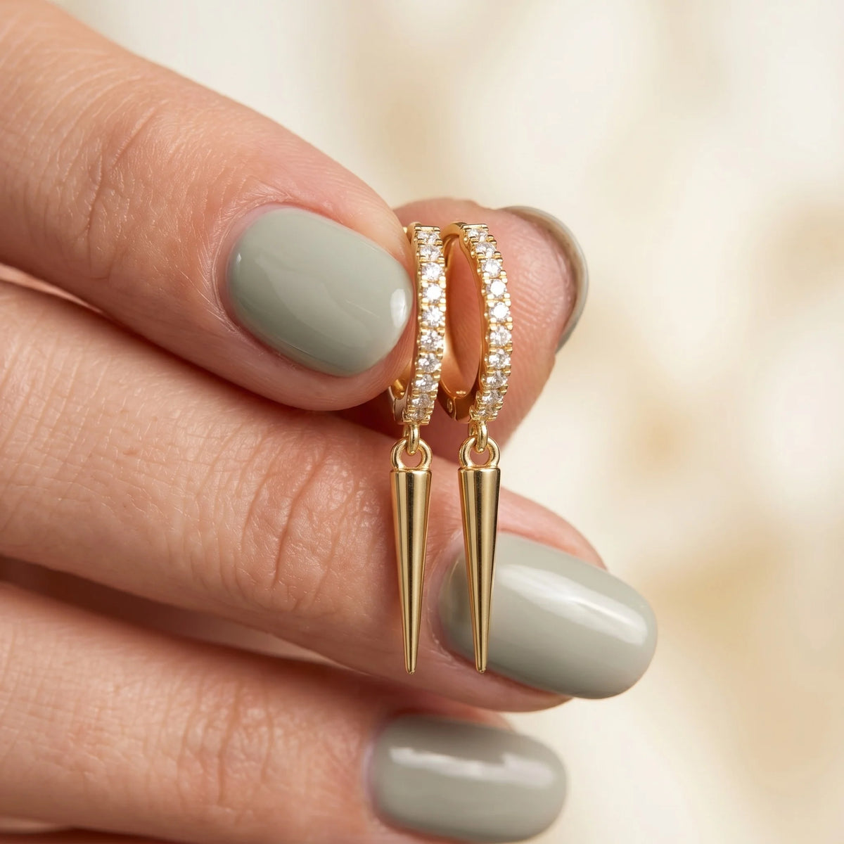 A person with sage green nail polish holding a pair of yellow gold diamond huggie hoop earrings featuring dangling cone spike charms.