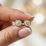 A pair of round diamond stud earring designs held between fingers, set in yellow gold with an octagonal bezel setting.