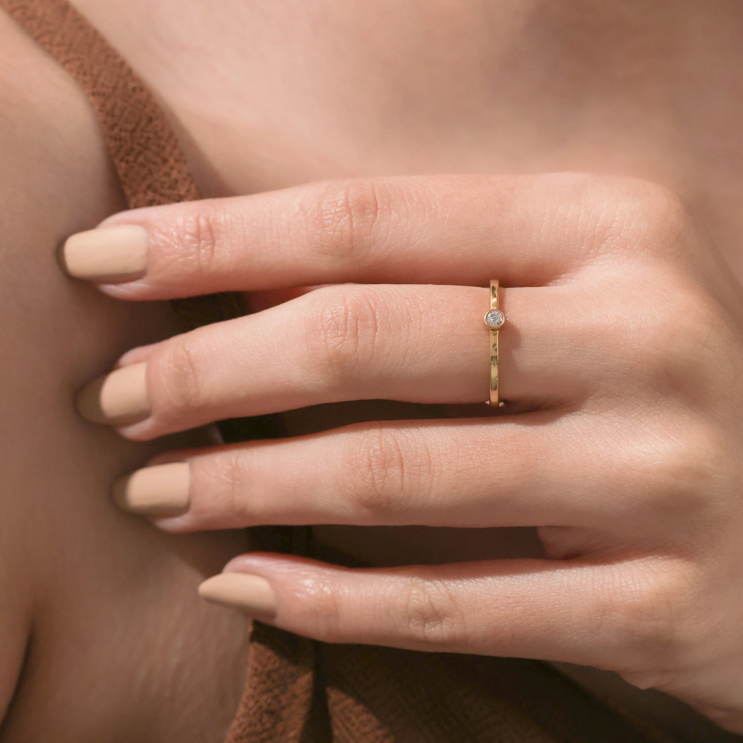 Close-up of a woman's hand wearing a thin gold bezel-set diamond ring, featuring neutral nude manicured nails.
