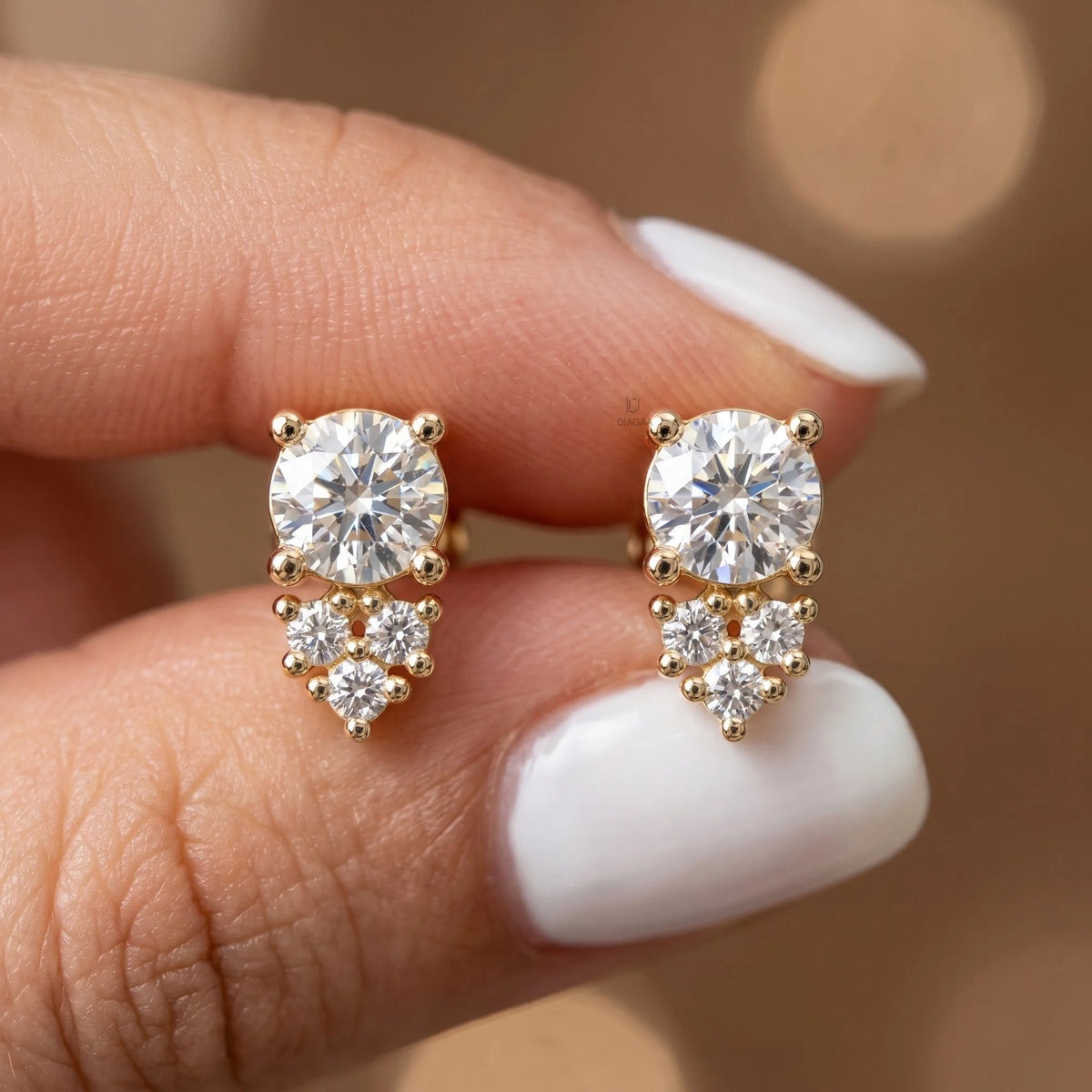 Pair of cluster round diamond stud earring pieces held between fingers, featuring a main round diamond with accent stones in yellow gold and a prong setting.