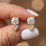 Pair of cluster round diamond stud earring pieces held between fingers, featuring a main round diamond with accent stones in yellow gold and a prong setting.
