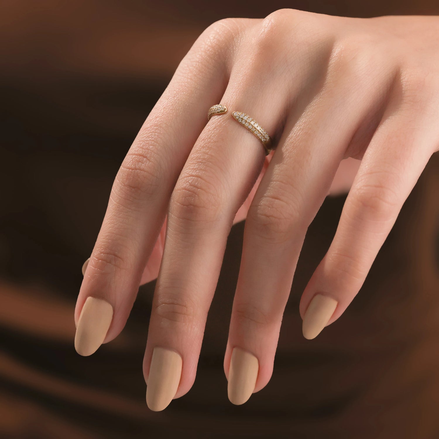 Delicate gold and diamond bypass ring modeled on a woman's middle finger. Shows a matte nude manicure and warm brown aesthetic, perfect for modern fine jewelry