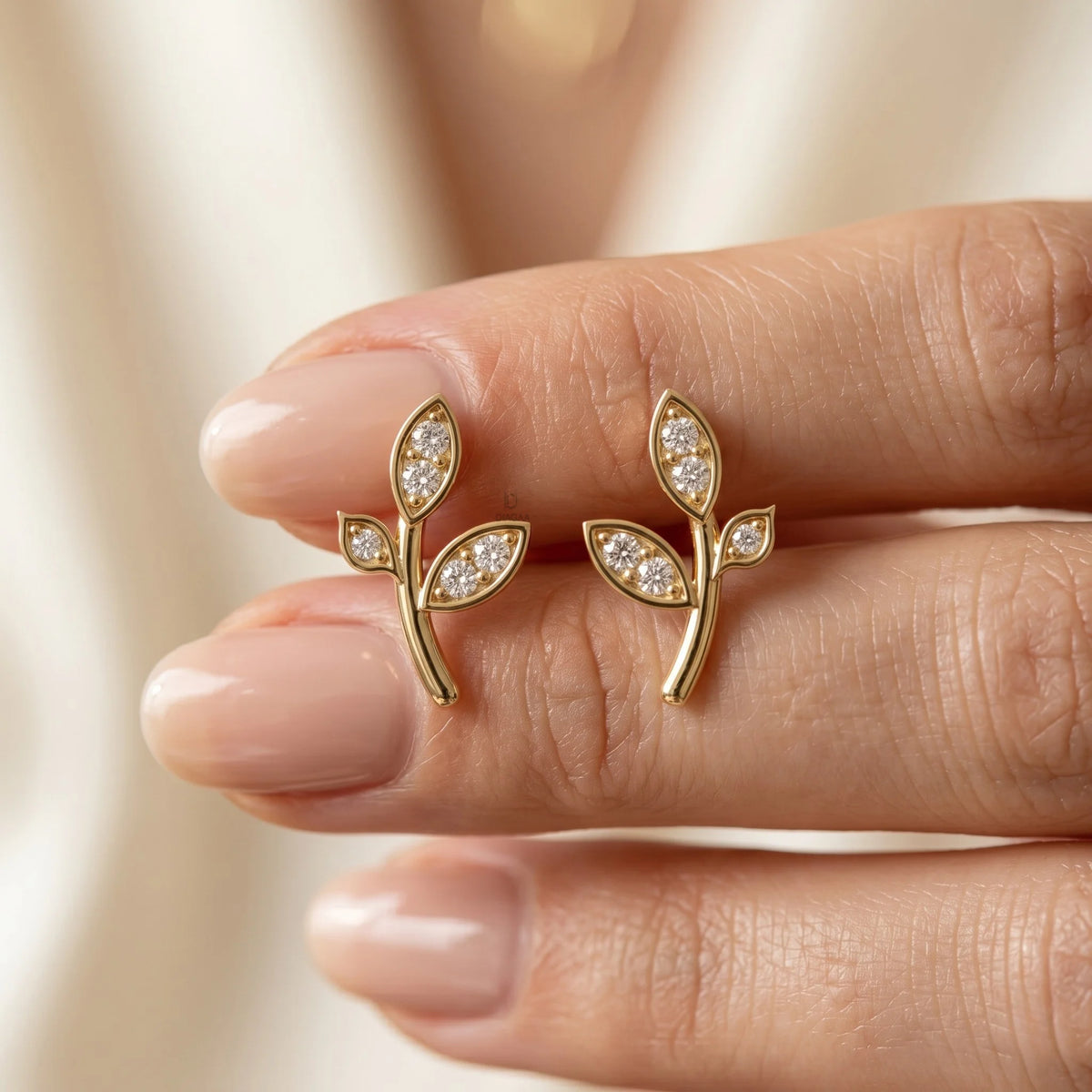 Pair of yellow gold leaf stud earrings held between fingers with a glossy manicure, showcasing the small size of the diamond branch jewelry.