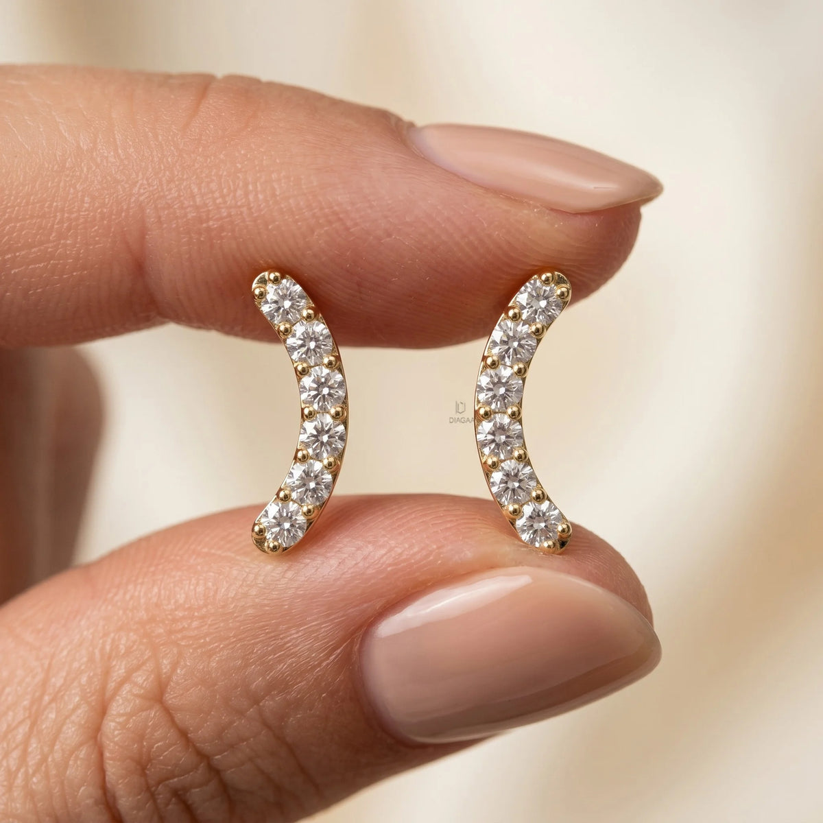 Fingers holding a pair of yellow gold curved round diamond stud earrings to show scale against a soft background