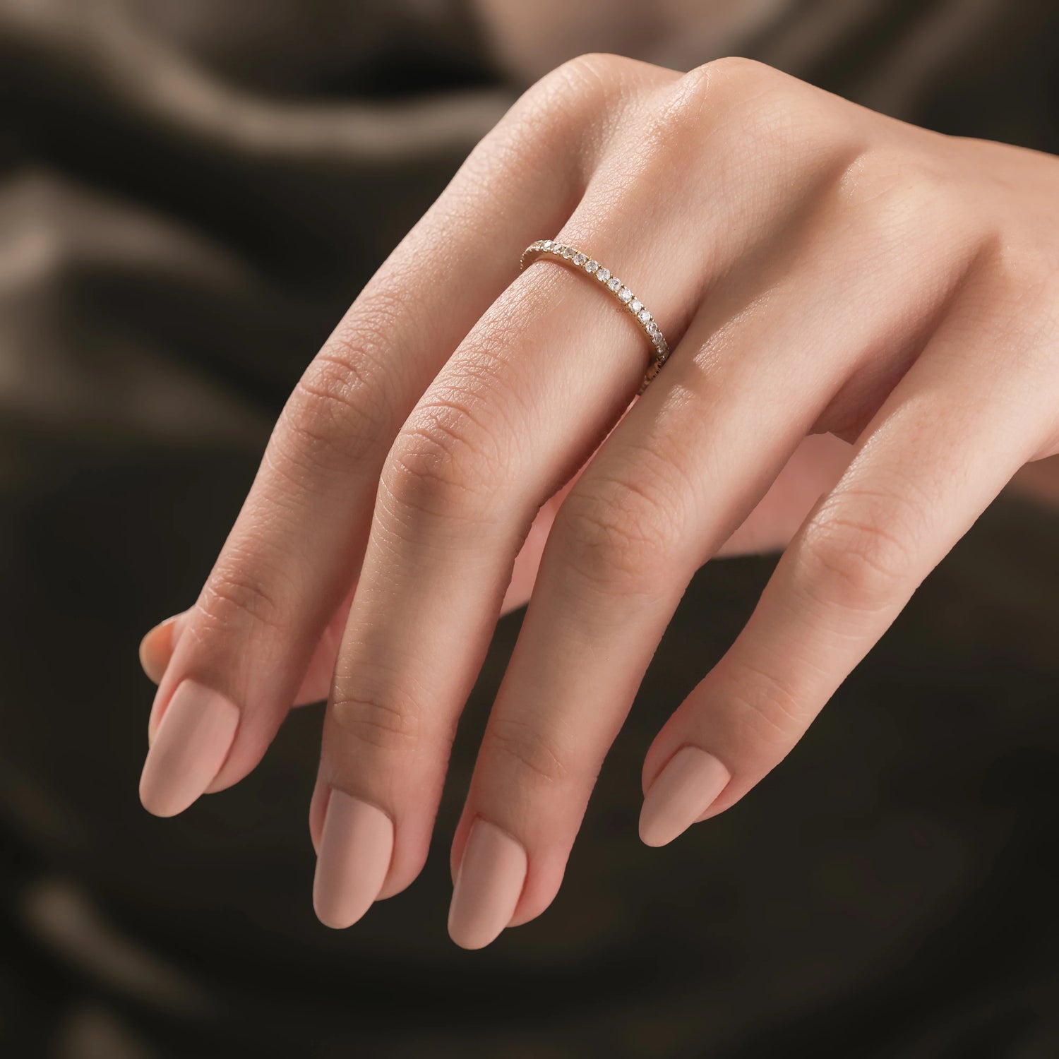 displays a minimalist yellow gold round diamond eternity stackable ring worn on a middle finger against a soft brown background. Perfect wedding band inspiration.