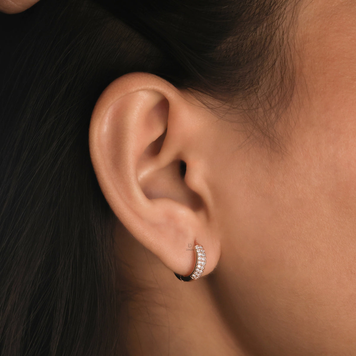 A woman wearing a wide gold huggie hoop earring encrusted with three rows of sparkling pavé set round diamonds in her earlobe piercing, touching her hair.
