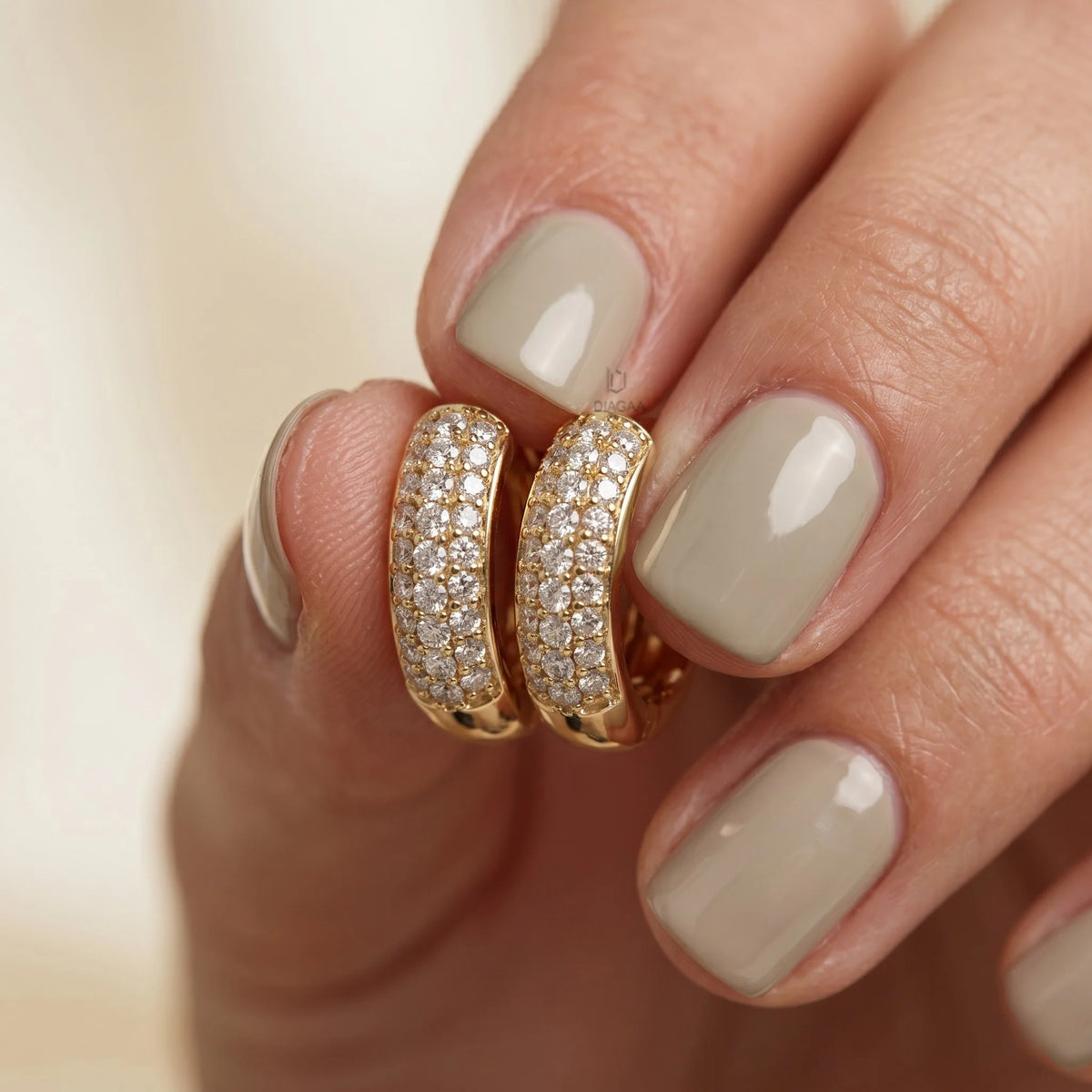 A hand with glossy sage green fingernails holding a pair of wide yellow gold huggie hoop earrings featuring three rows of brilliant pavé set round diamonds hoops earring.