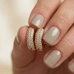 A hand with glossy sage green fingernails holding a pair of wide yellow gold huggie hoop earrings featuring three rows of brilliant pavé set round diamonds hoops earring.