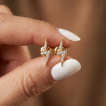 A person with white manicured nails holding a pair of dainty yellow gold lightning bolt stud earrings encrusted with clear pave diamonds.