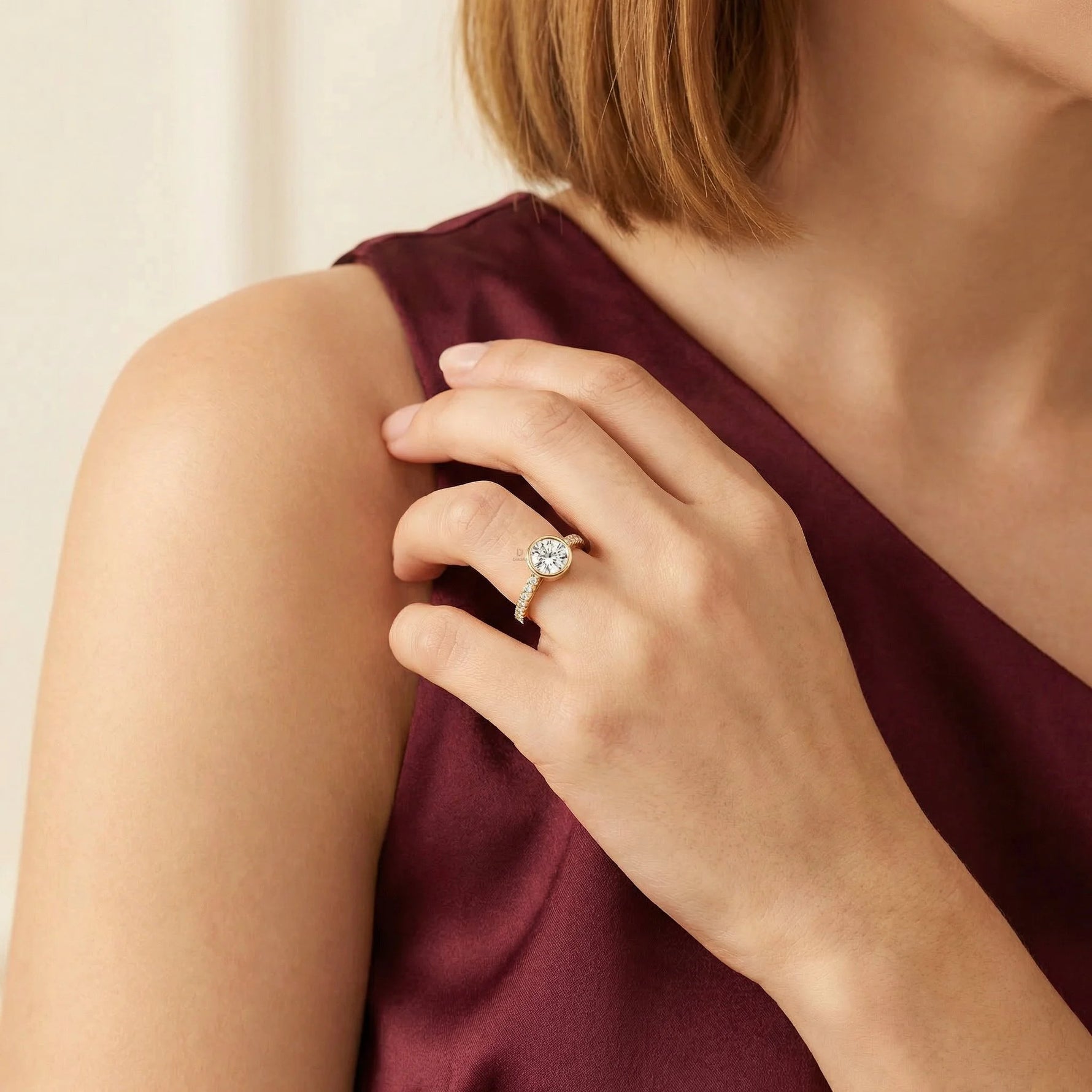 Close-up of a yellow gold round diamond engagement ring with a modern bezel setting and shimmering pavé band, styled on a woman's hand wearing a burgundy top.