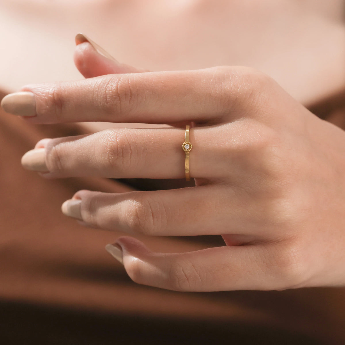 Close up of a minimalist gold promise ring with a thin band and hexagon bezel gemstone on a finger.