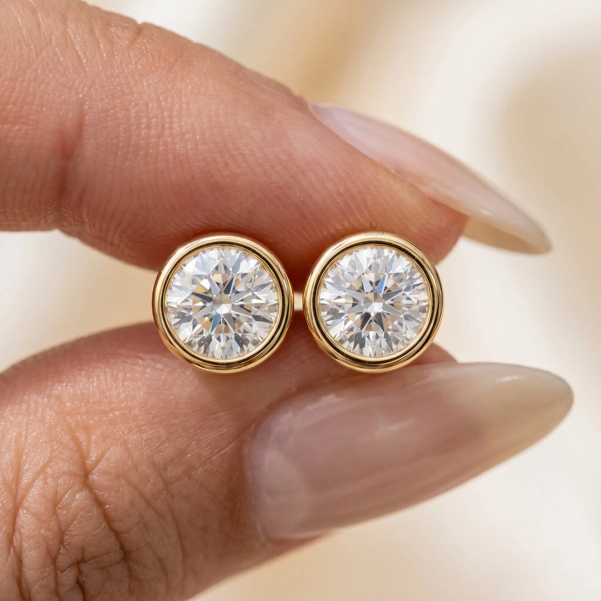 Close-up of fingers holding a pair of yellow gold round bezel-set diamond stud earrings to show size and brilliance.